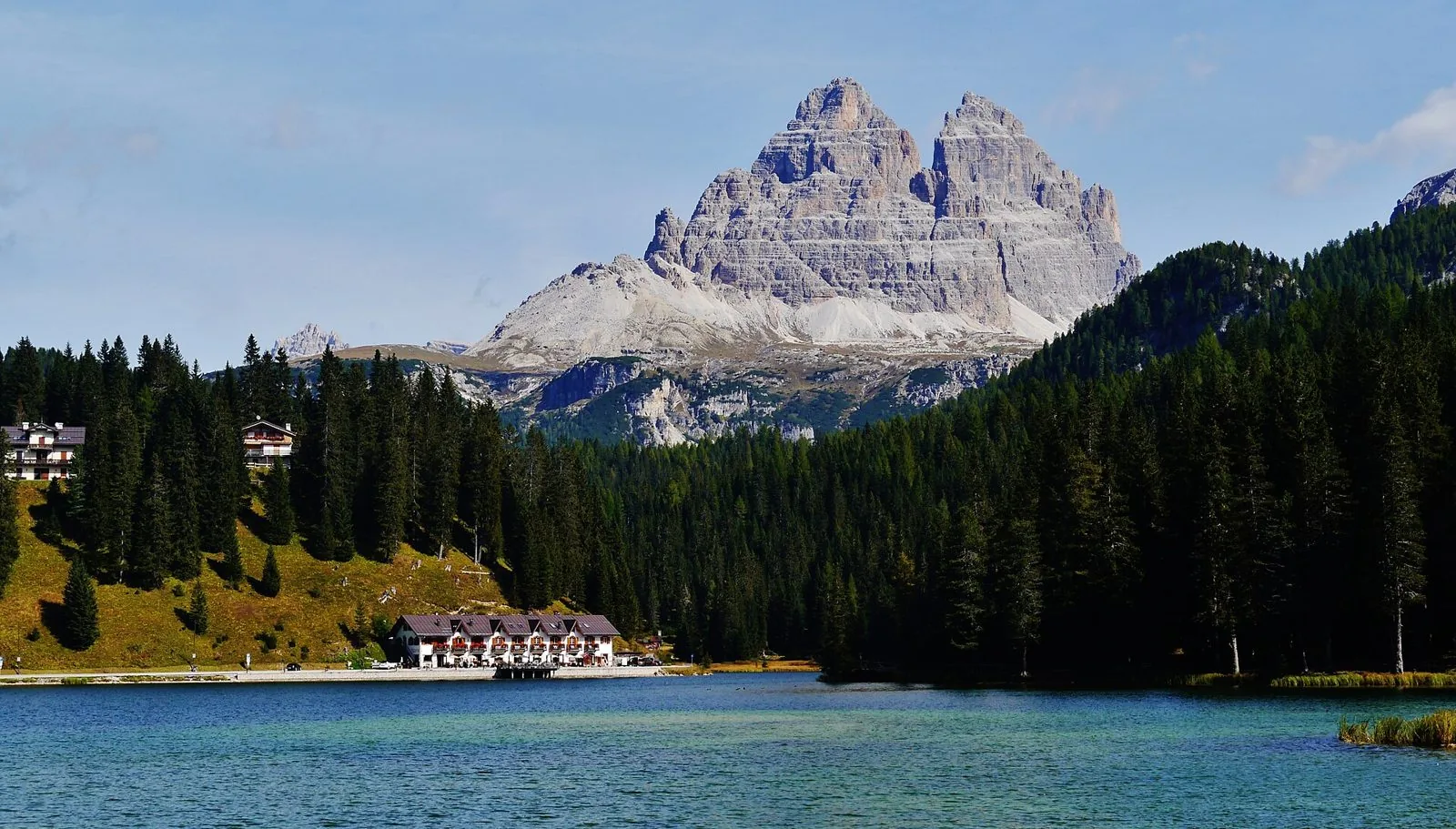 Il lago di Misurina con acqua turchese, un edificio sulla riva e boschi di conifere, con il versante sud delle Tre Cime di Lavaredo sullo sfondo
