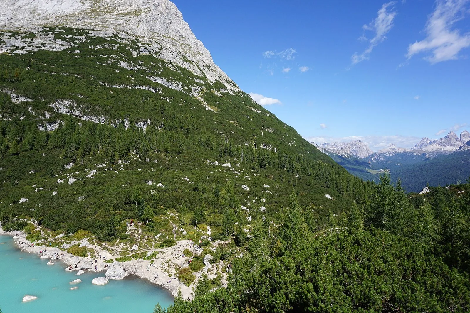 Le acque turchesi del lago del Sorapiss viste dall'alto, con la riva sassosa, i pendii boscosi del Sorapiss e le Tre Cime di Lavaredo lontane all'orizzonte