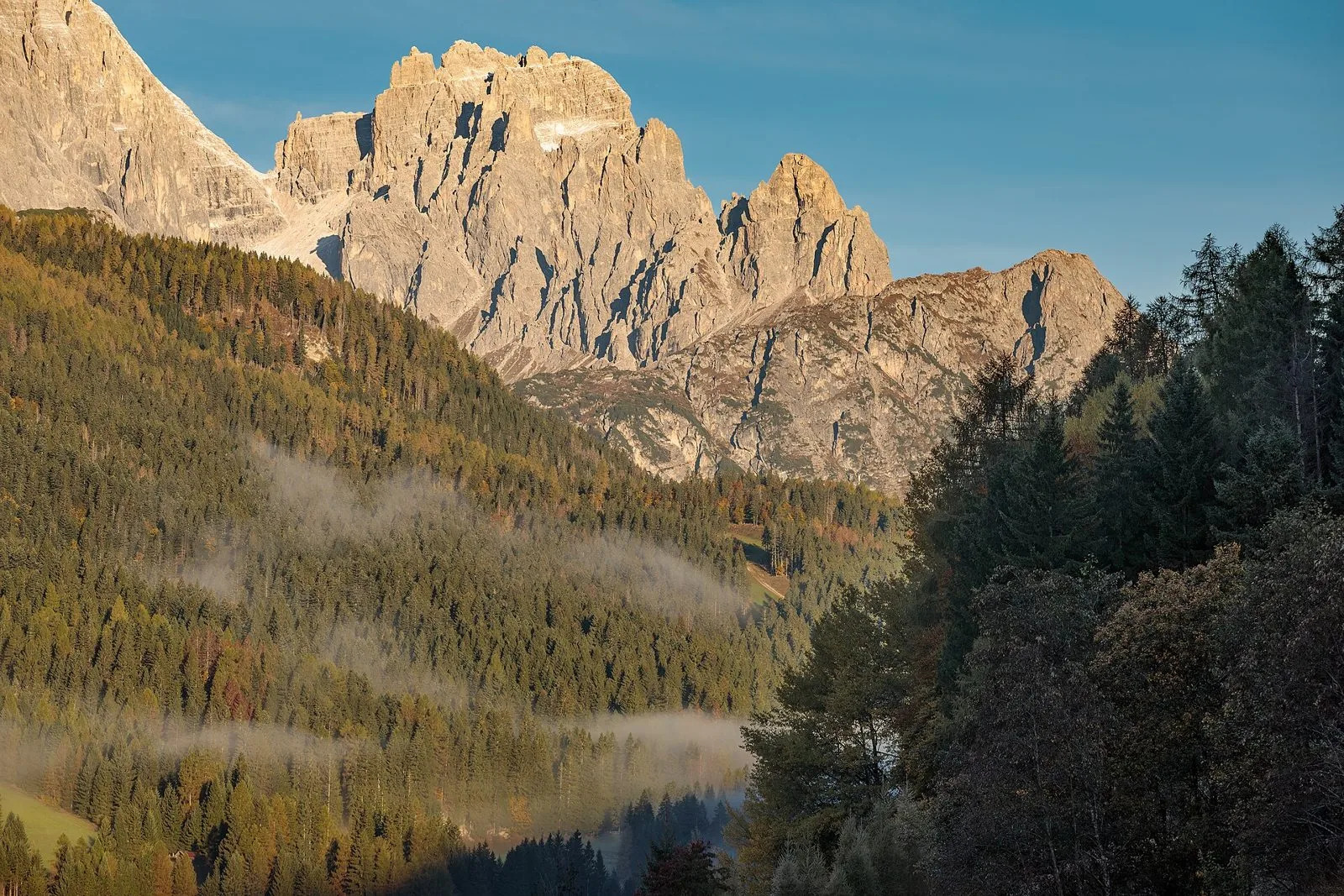 Pareti rocciose delle Dolomiti di Sesto illuminate dal sole sopra boschi di abeti e larici autunnali, con nebbie basse nella valle vicino a Dosoledo