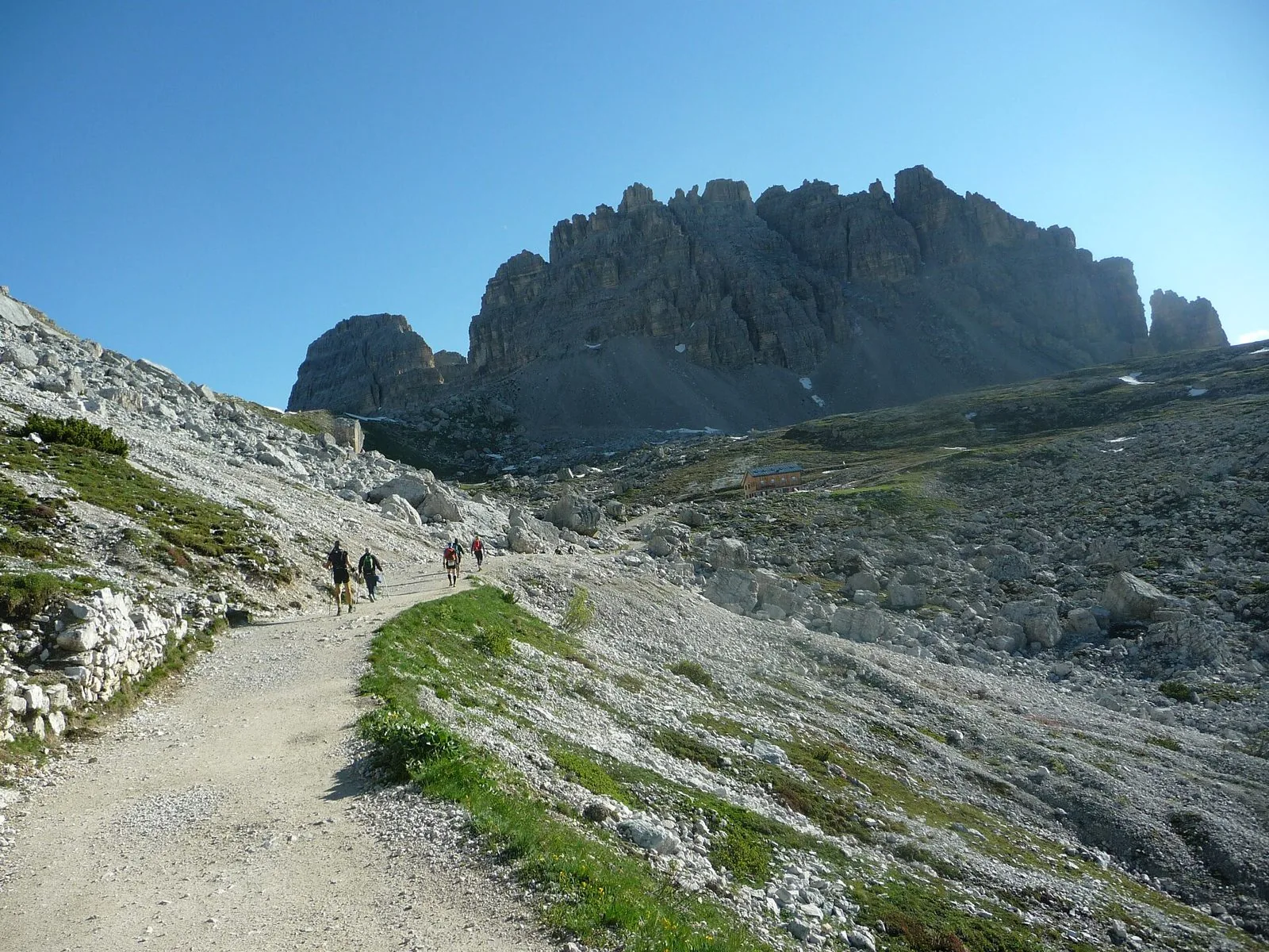 Sentiero sterrato che sale verso la Forcella Lavaredo, con alcuni escursionisti di spalle, un rifugio sullo sfondo e alte pareti dolomitiche sotto il cielo azzurro